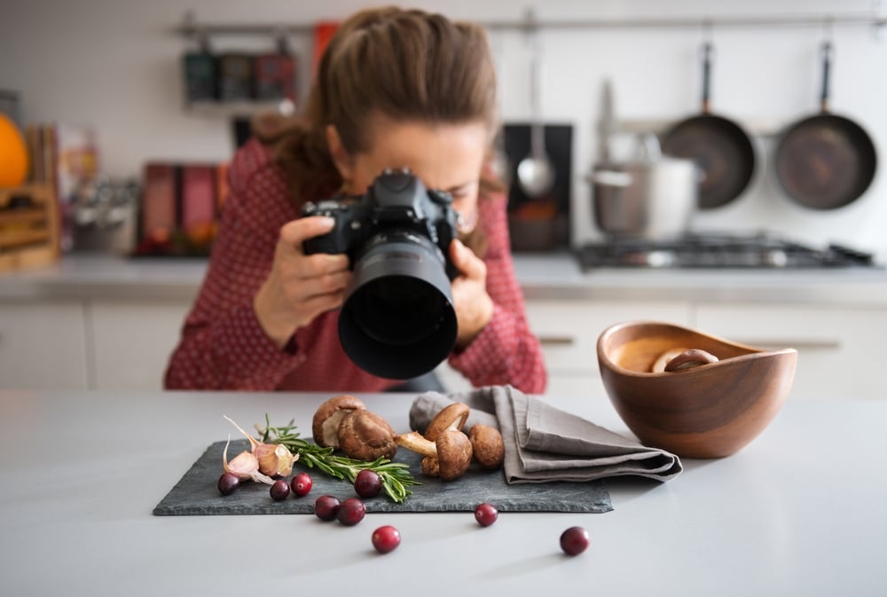 A woman food photographer in the background leans down to take a close-up, in a modern kitchen, of autumn fruits and vegetables - mushrooms, garlic, rosemary, and cranberries.