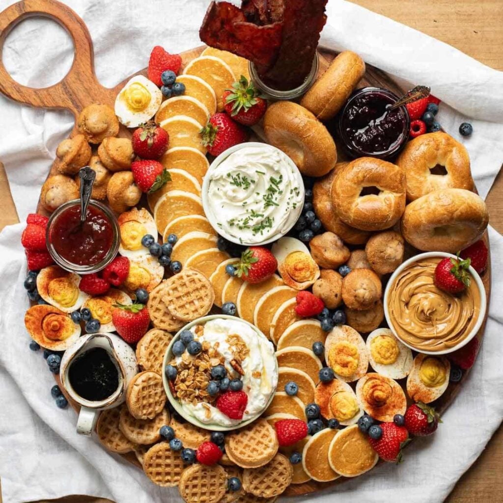 Breakfast charcuterie board with mini pancakes, waffles, bagels, bacon, deviled eggs and berries