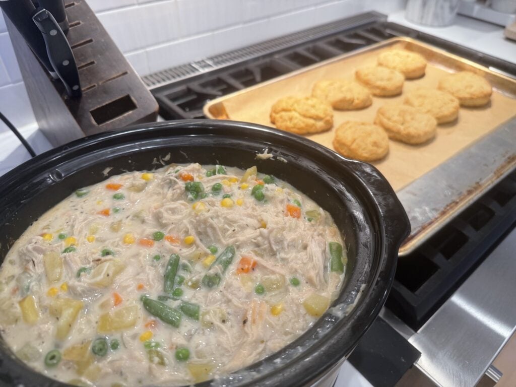 Finished chicken pot pie filling in the crockpot next to a tray of baked biscuits